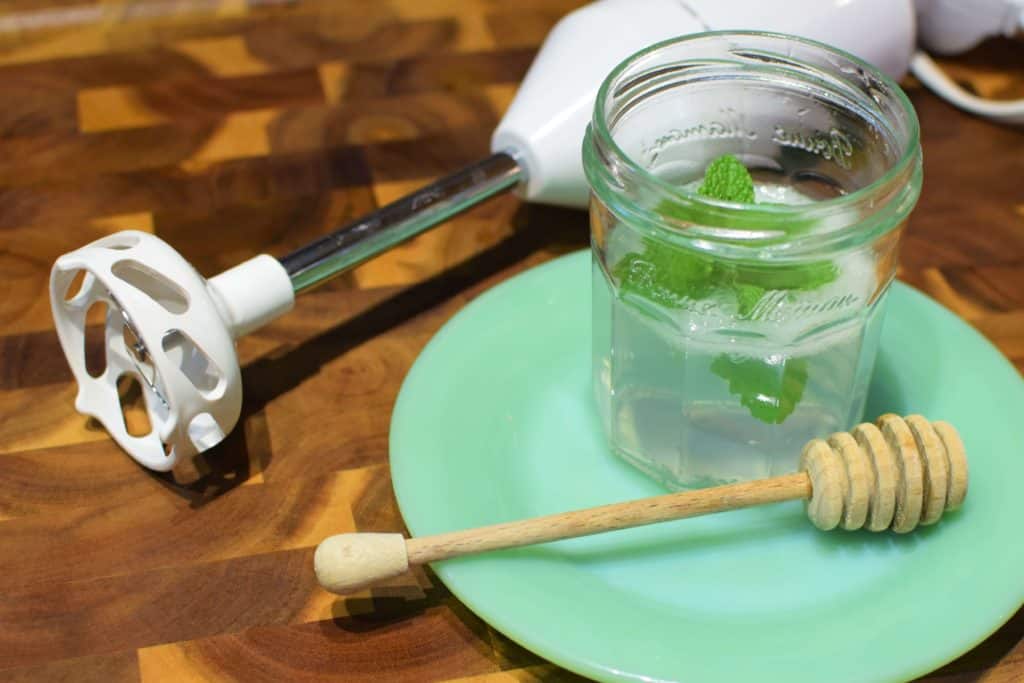 A clear glass jar full of freshly made aloe vera gel sitting on a jade green milk glass plate. Next to the plate is a white immersion blender. On the plate is a wooden honey-dipper.