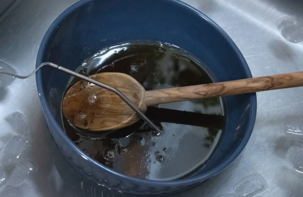 A bowl of hot maple syrup resting in an ice bath in the sink. A wooden spoon and a digital thermometer electrode are sticking out of the syrup.