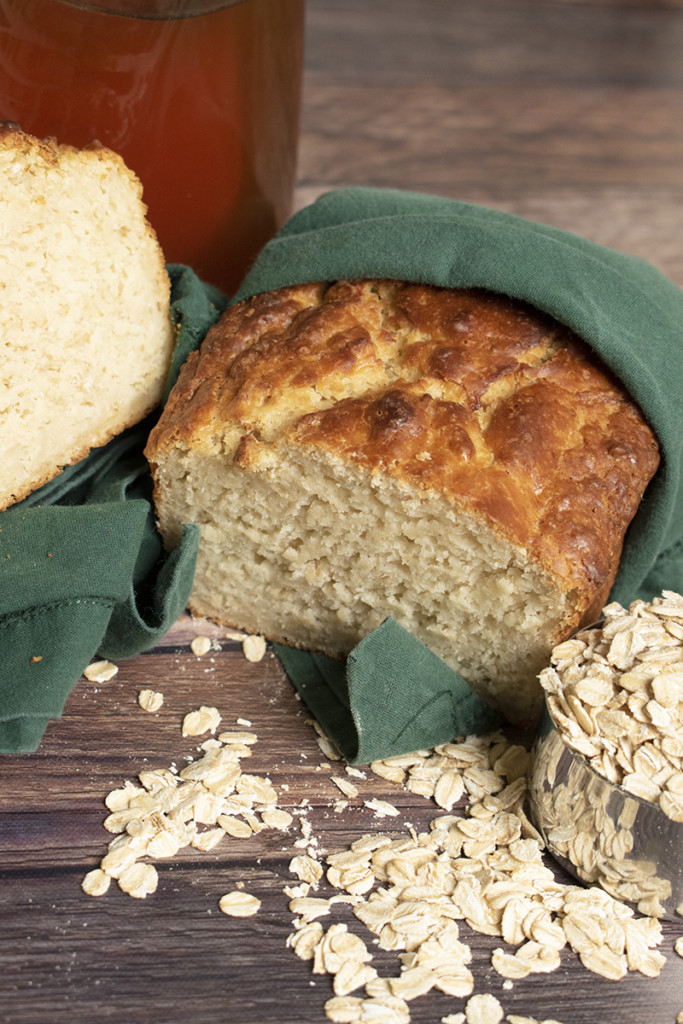 A loaf of bread is wrapped in a towel; it sits next to rolled oats in a measuring cup.