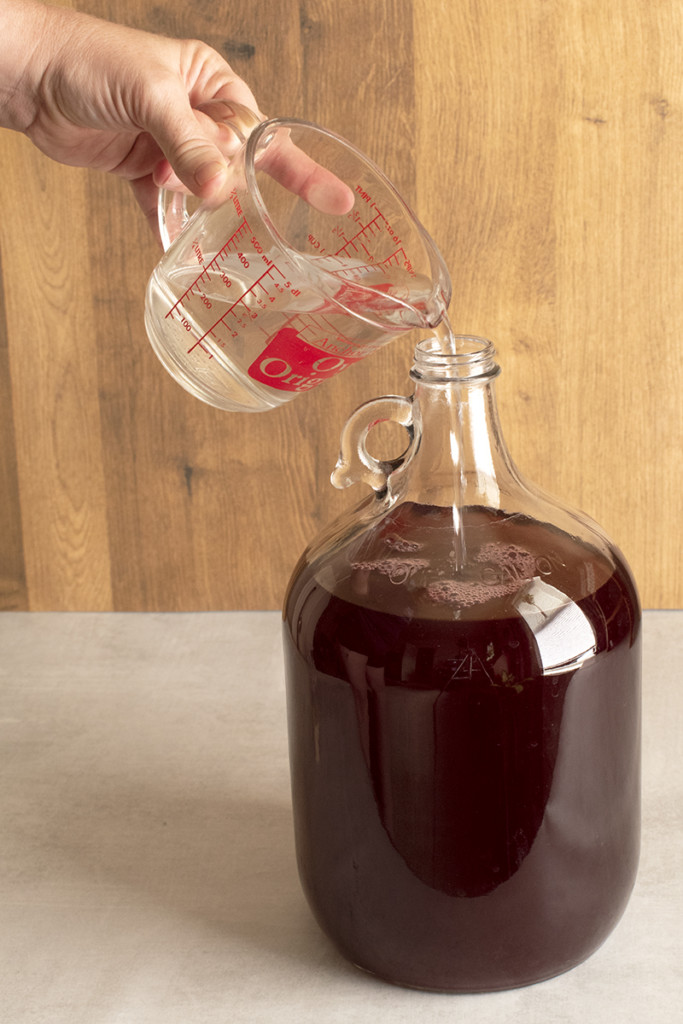 a hand pours water into a carboy of blueberry basil mead