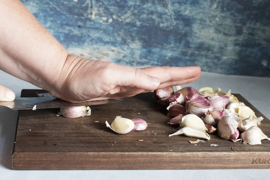 A hand using the flat of a knife blade to remove the paper skin from a clove of garlic.