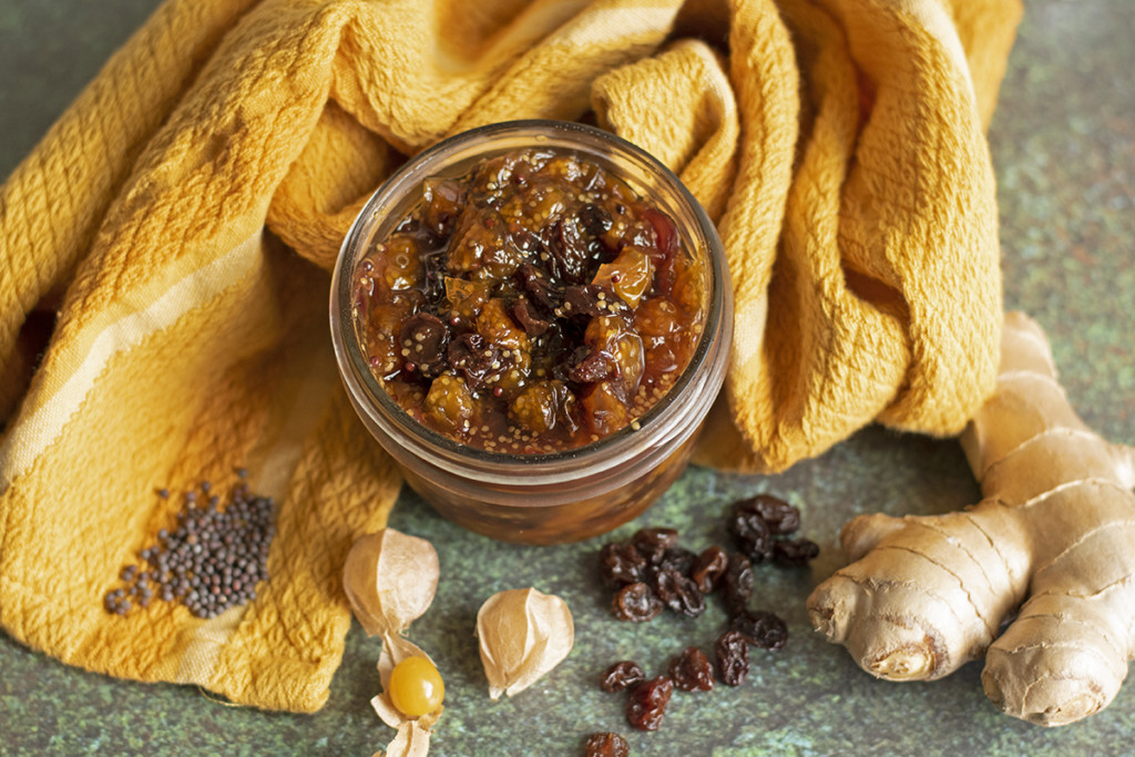 A jar of ground cherry chutney is surrounded by ginger, raisins and mustard seeds