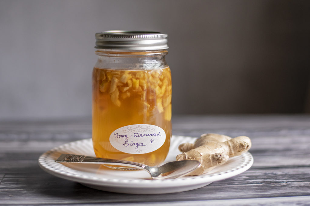 A jar of honey-fermented ginger is sitting on a white plate next to a spoon and a piece of whole ginger.