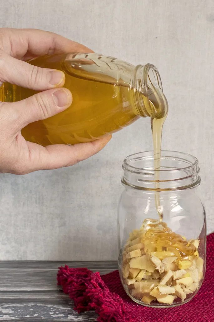 Two hands pour honey from a jar into a mason jar filled halfway with chopped, fresh ginger.
