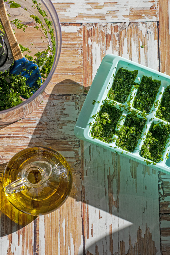 A food processor bowl containing minced basil leaves, and a cruet of olive oil sit next to an ice cube tray containing minced basil leaves and olive oil to be frozen.