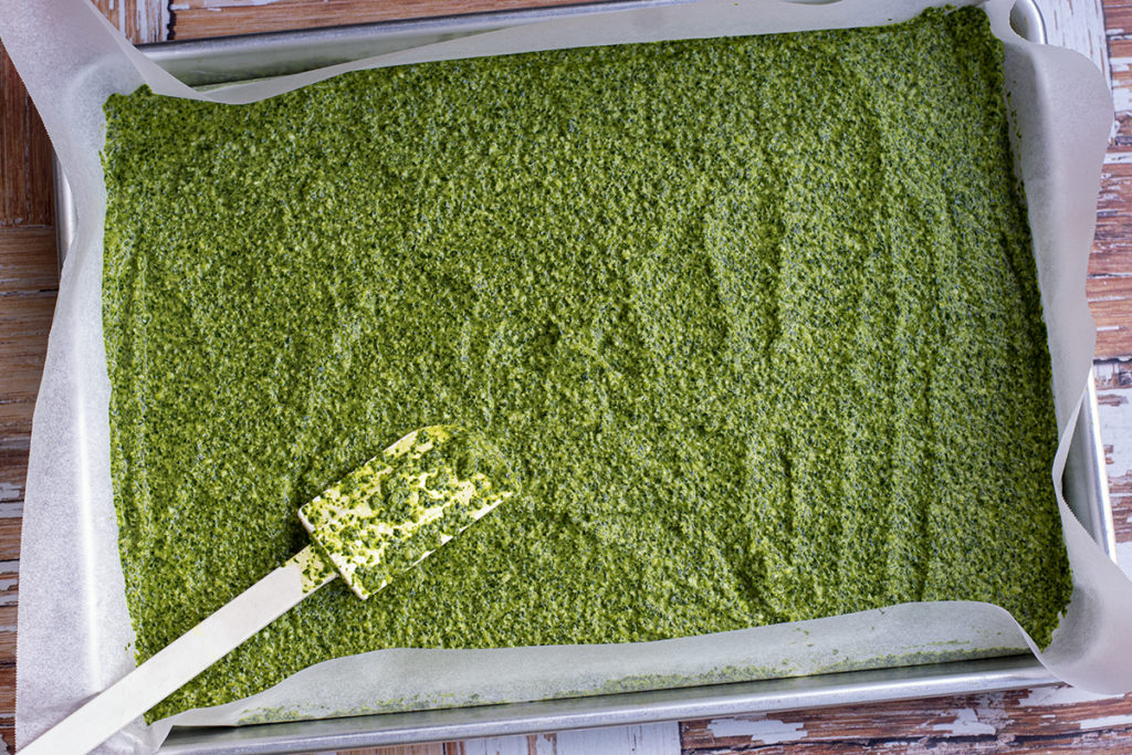 Pesto is spread in a thin layer on a parchment-lined baking sheet with a rubber spatula. 