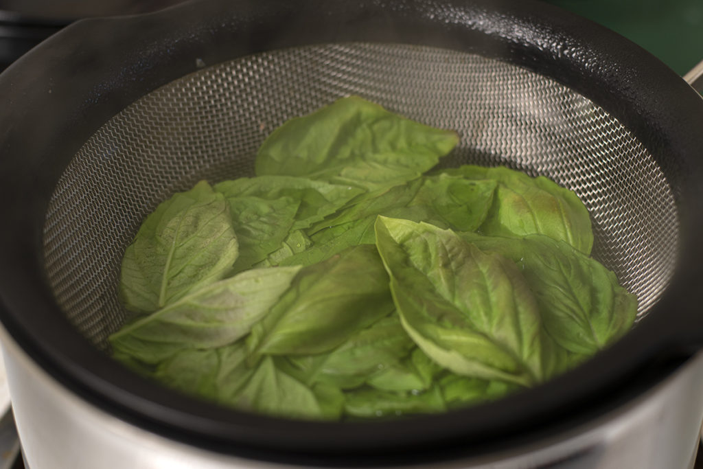 Basil leaves in a mesh strainer being lowered into boiling water. 