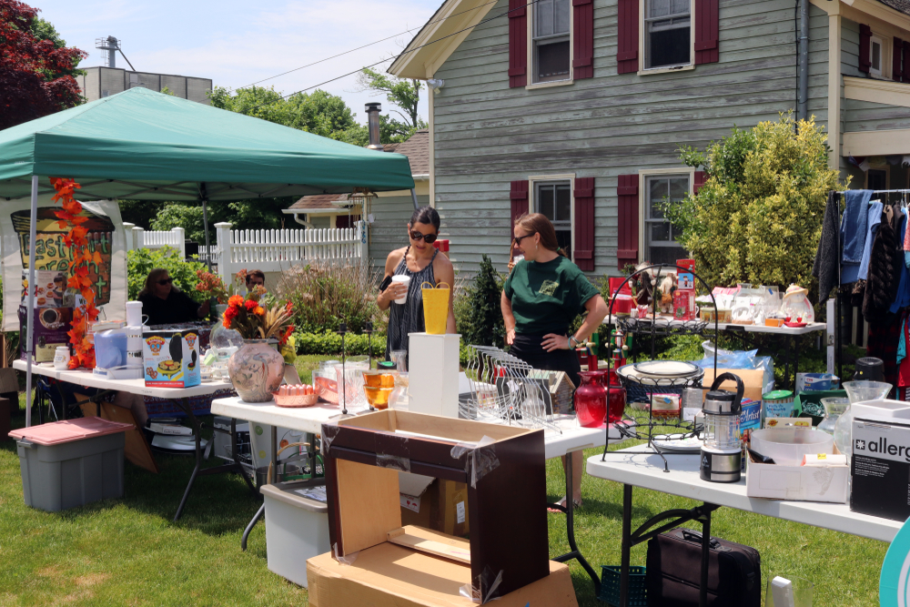 Two women talk at a yard sale. Tables in a yard with household items for sale.