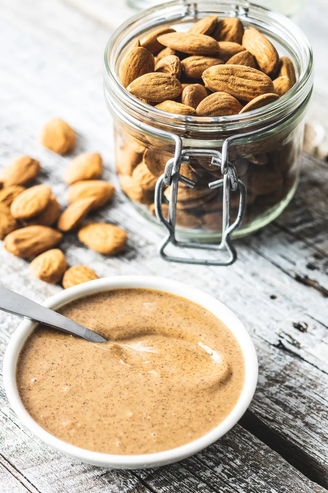 A small dish of almond butter with a spoon in it sits next to a jar of almonds.