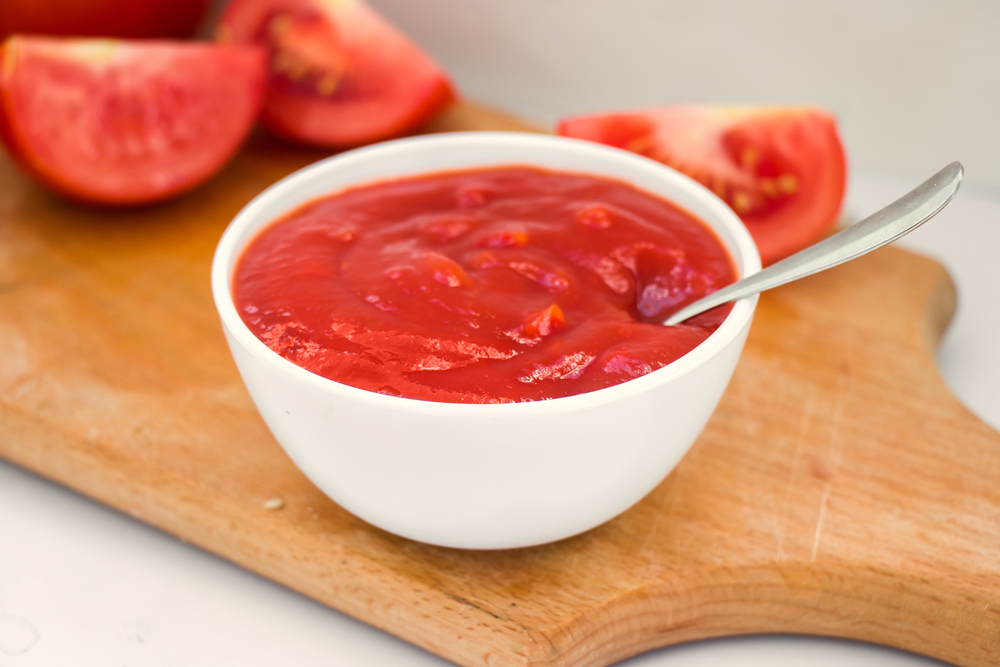 A small dish of homemade tomato ketchup sits on a cutting board with sliced tomatoes next to it.