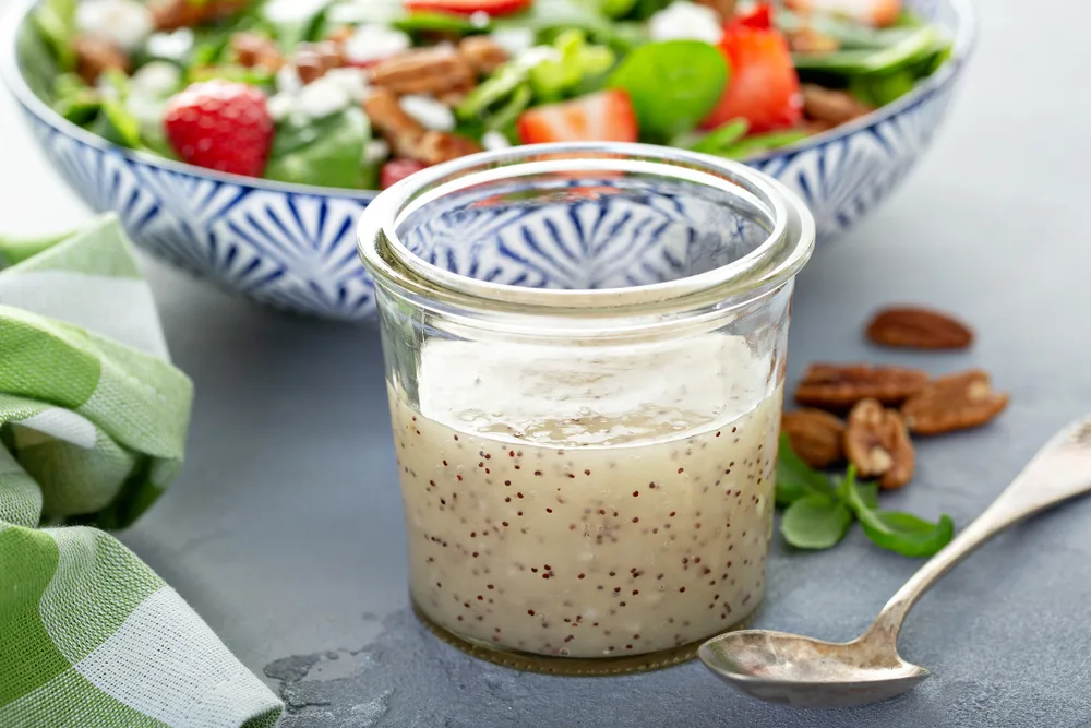 A jar of homemade salad dressing sits on a table with a spoon and a salad.