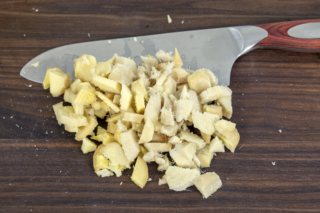 A cutting board with 1/2" chunks of roughly chopped ginger with the knife sitting behind it.
