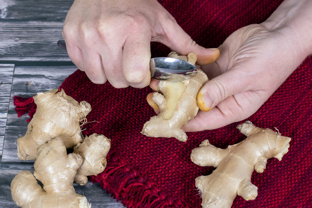 A pair of hands are shown using the side of a spoon to peel fresh ginger.