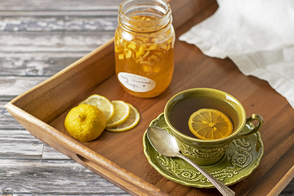 A jar of honey-fermented ginger, lemon slices, a tea towel, and a cup of tea and saucer are all arranged on a tea tray.