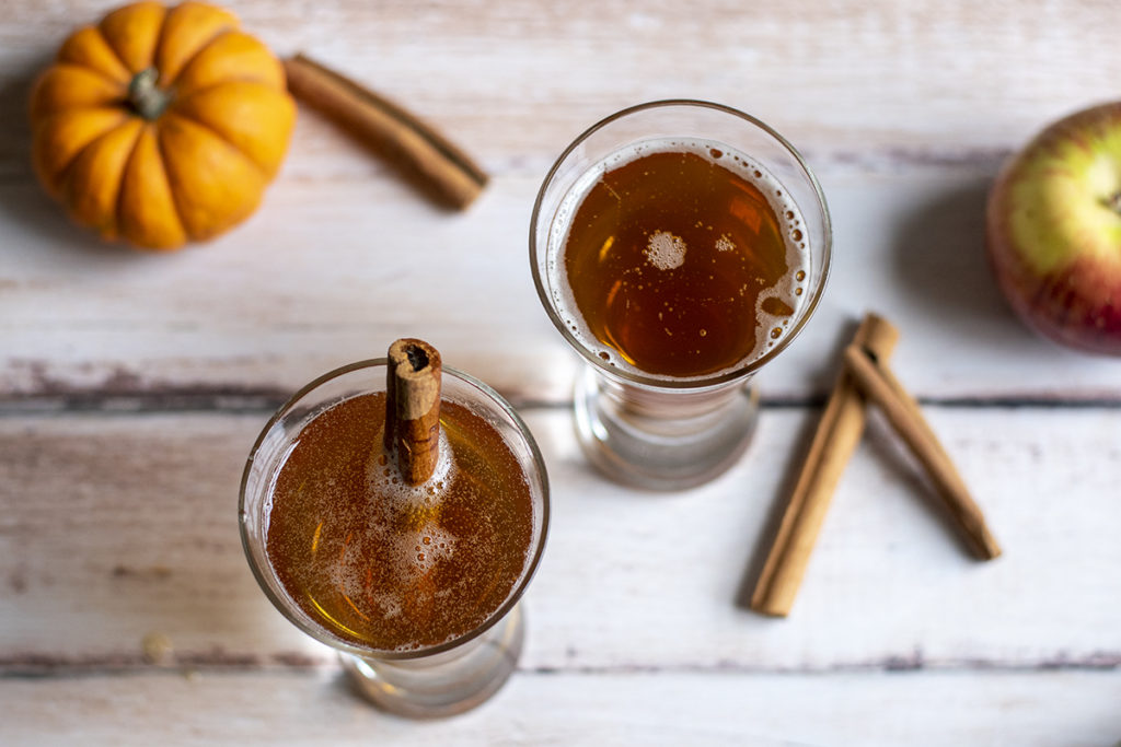 Two glasses of spiced pumpkin cider are set next to a tiny pumpkin and an apple.