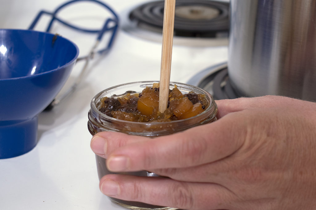 A hand is shown using a chopstick to remove air bubbles from the hot pumpkin chutney.