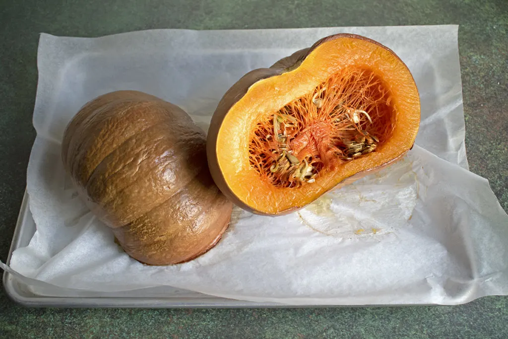 Two halves of a roasted pumpkin lay face down on a parchment paper-lined baking sheet.