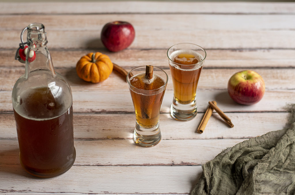 Two glasses of spiced pumpkin cider are sitting next to the bottle of cider on a wooden floor with apples, cinnamon sticks and tiny pumpkins.