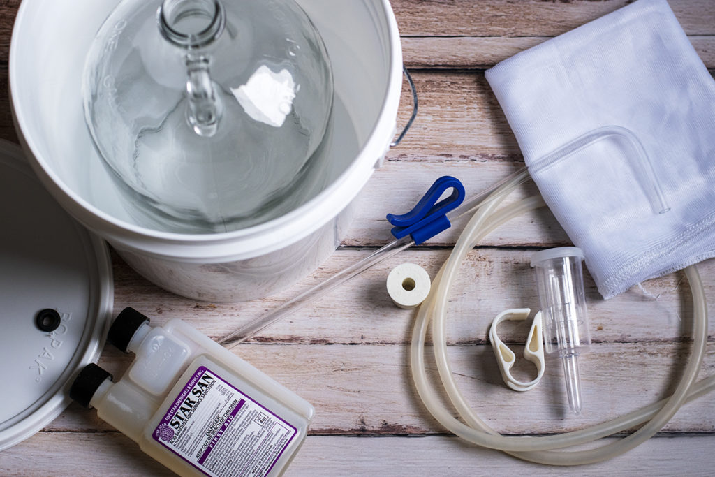 Homebrewing equipment pieces are laid out on a wooden floor: a plastic bucket with lid, a glass jug, sanitizer, a racking cane, a nylon straining bag, a length of silicon tubing, a tubing clamp and a 3-piece airlock and rubber stopper.