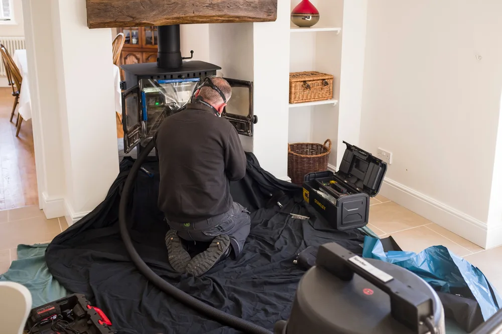 A professional kneels on a cloth covered floor to clean and inspect a woodstove.
