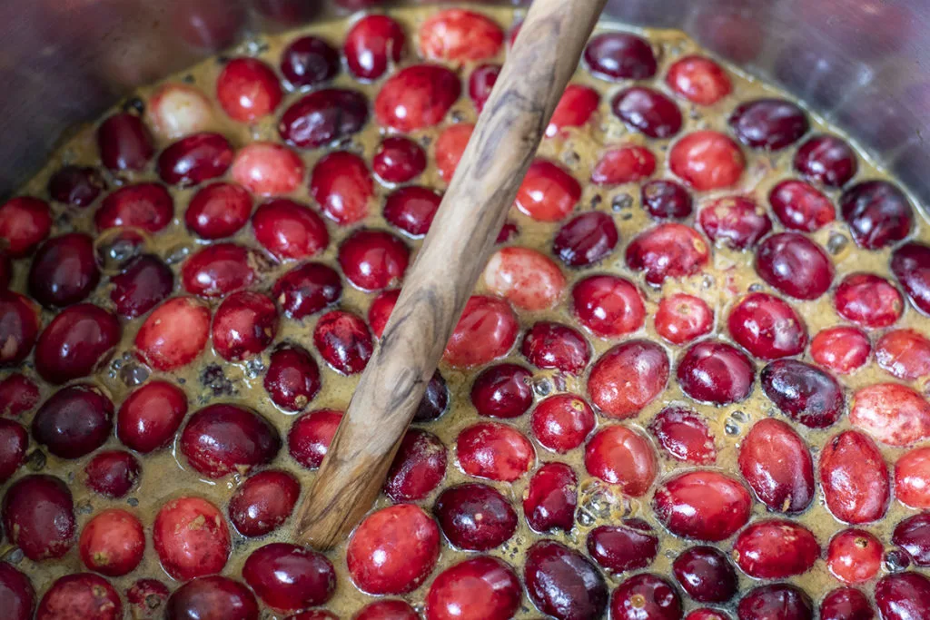 Cranberries and cider are being simmered together in a large stockpot with a wooden spoon in it.