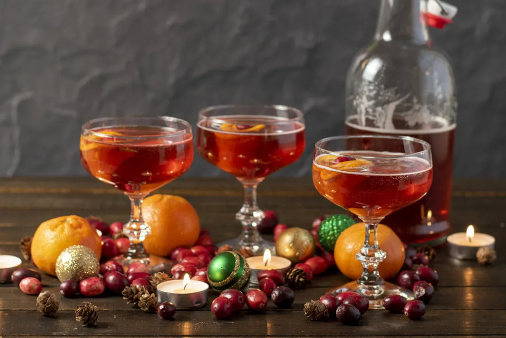 Three coupe glasses filled with pink Christmas cider in front of a bottle of cider. There are cranberries, tiny pinecones, candles, clementines and Christmas baubles on the table around the glasses.