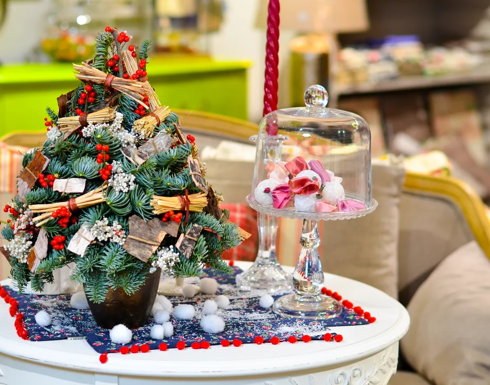 A tiny tabletop Christmas tree decorated with ornaments on a table  next to a dish full of candy and a candlestick.
