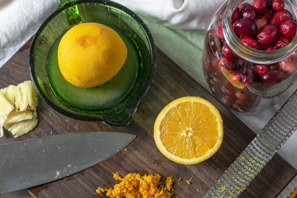 A cutting board is shown with an orange half, orange zest, a smashed piece of garlic, a juicer with an orange half, a Microplane zester and a chef's knife. Next to the cutting board is a jar of cranberries.