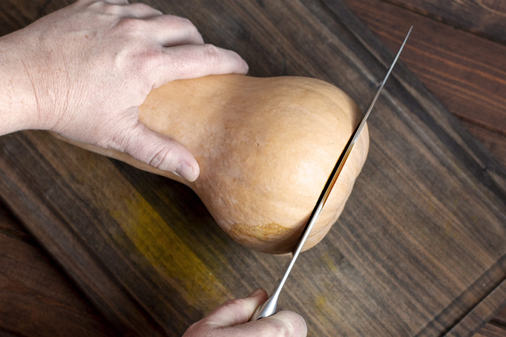 Hands are shown slicing the end off of a butternut squash on a cutting board.