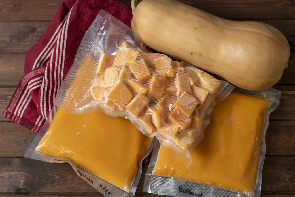 Bags of frozen butternut squash, pureed and cubed next to a whole butternut squash on a burgundy tea towel.