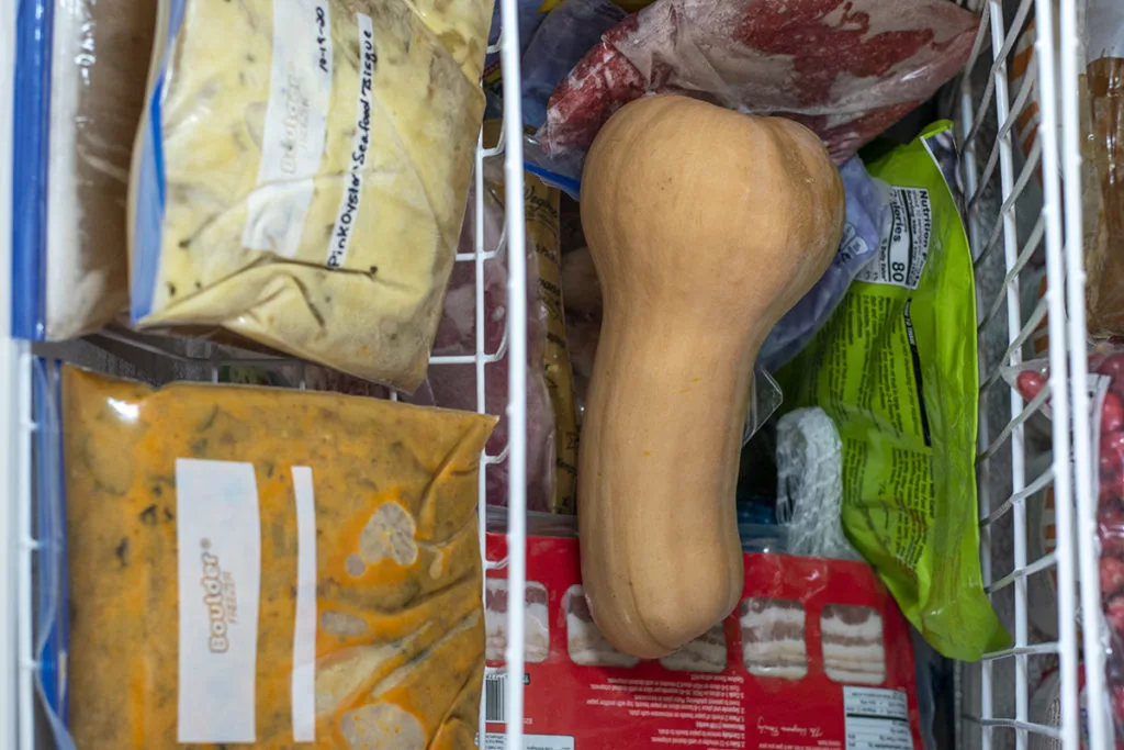 A whole frozen butternut squash in a freezer surrounded by packages of frozen food.