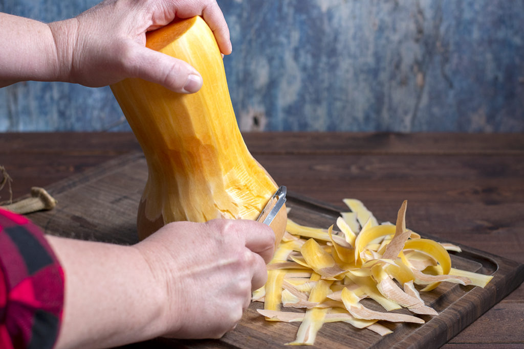 Hands are showing peeling the skin from a butternut squash. There are peels on a cutting board next to the squash.