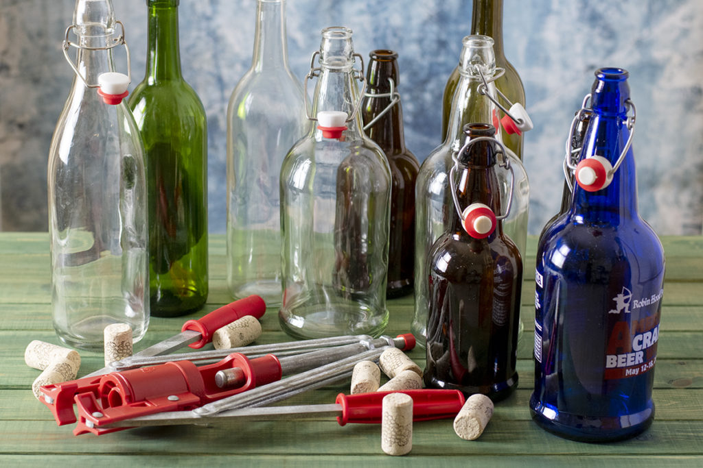 An assortment of empty wine bottles and swing-top bottles next to a wine corker with corks.
