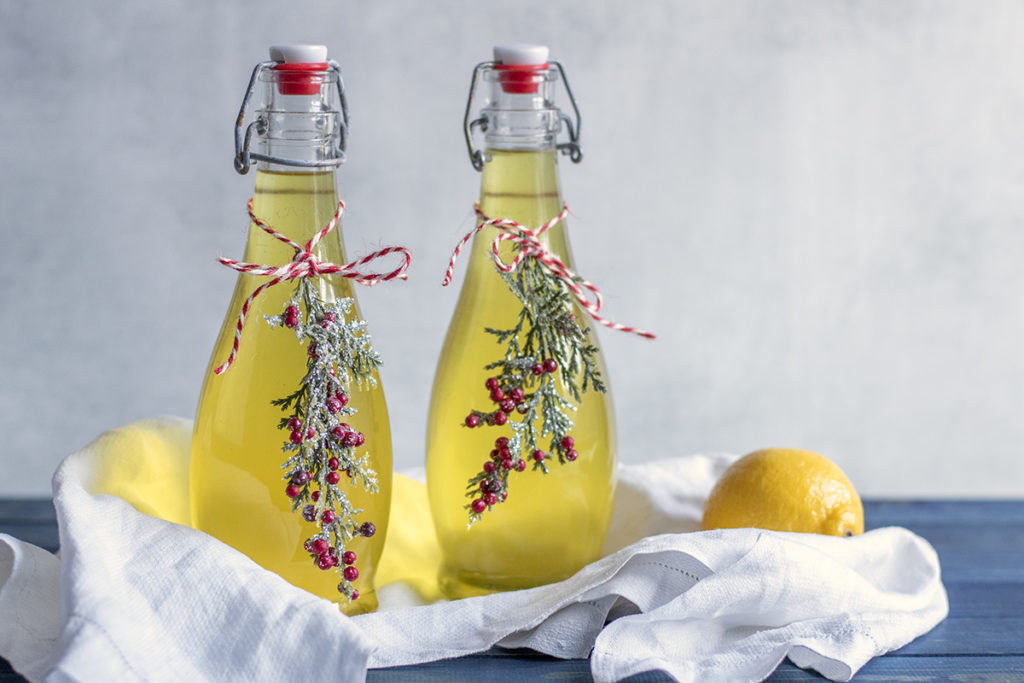 Two bottles of homemade limoncello decorated with holly sprigs and twine. The bottles sit on a wooden surface with a tea towel and a lemon.