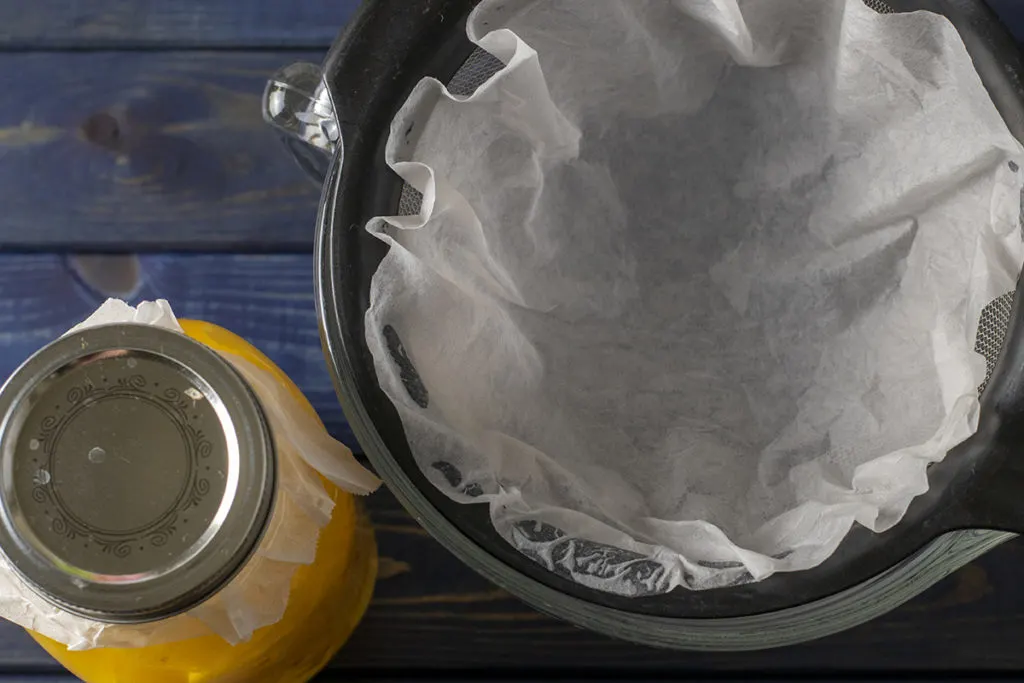 A wood surface with a jar of lemon peels in vodka and a bowl with a mesh strainer in it. There is a coffee filter in the mesh strainer.