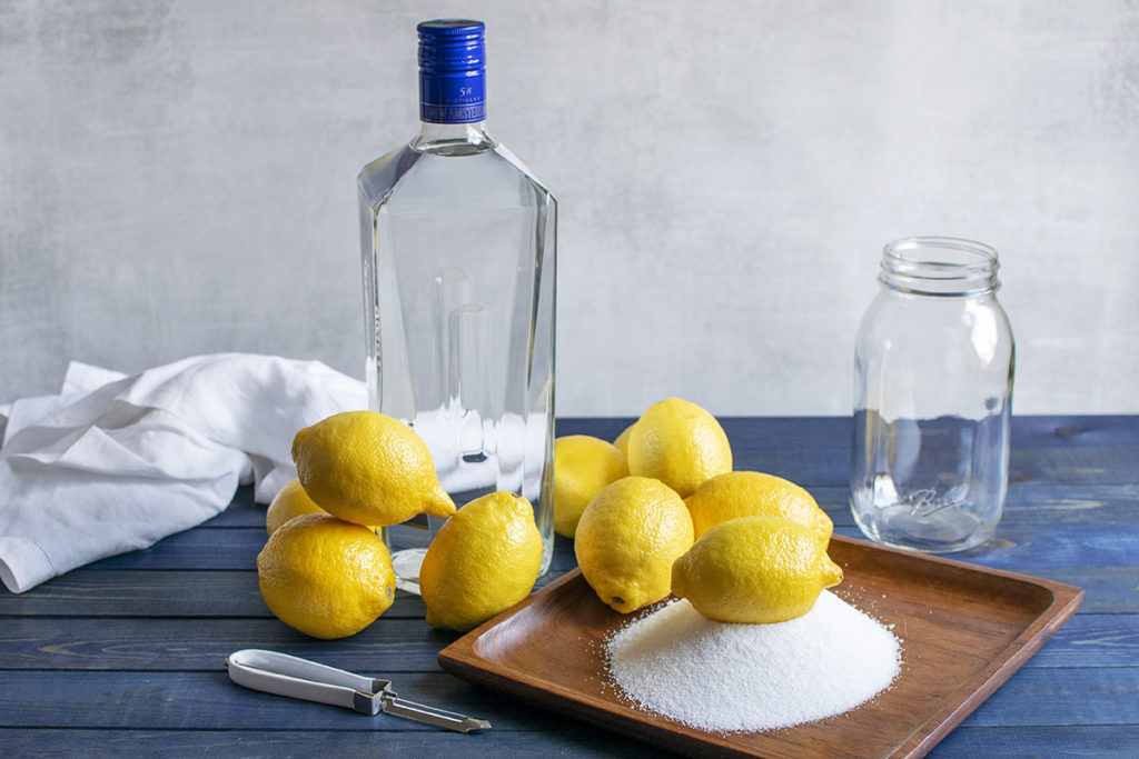 Limoncello ingredients are arranged on a table top: lemons, sugar and a bottle of vodka. A jar, a tea towel and a vegetable peeler are laid out nearby.