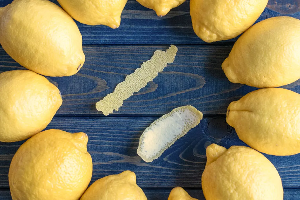 Two strips of lemon peel are encircled by whole lemons. One peel is very thin, the other is thicker with the white pith attached to it.