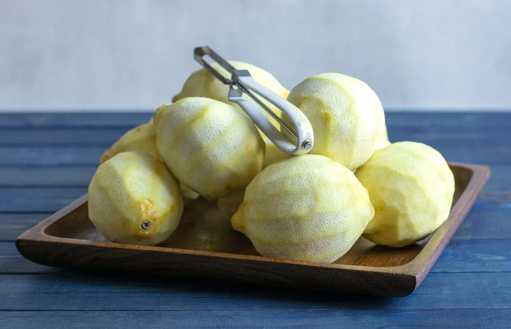 A pile of peeled lemons on a teak dish with a vegetable peeler on top of the lemons.