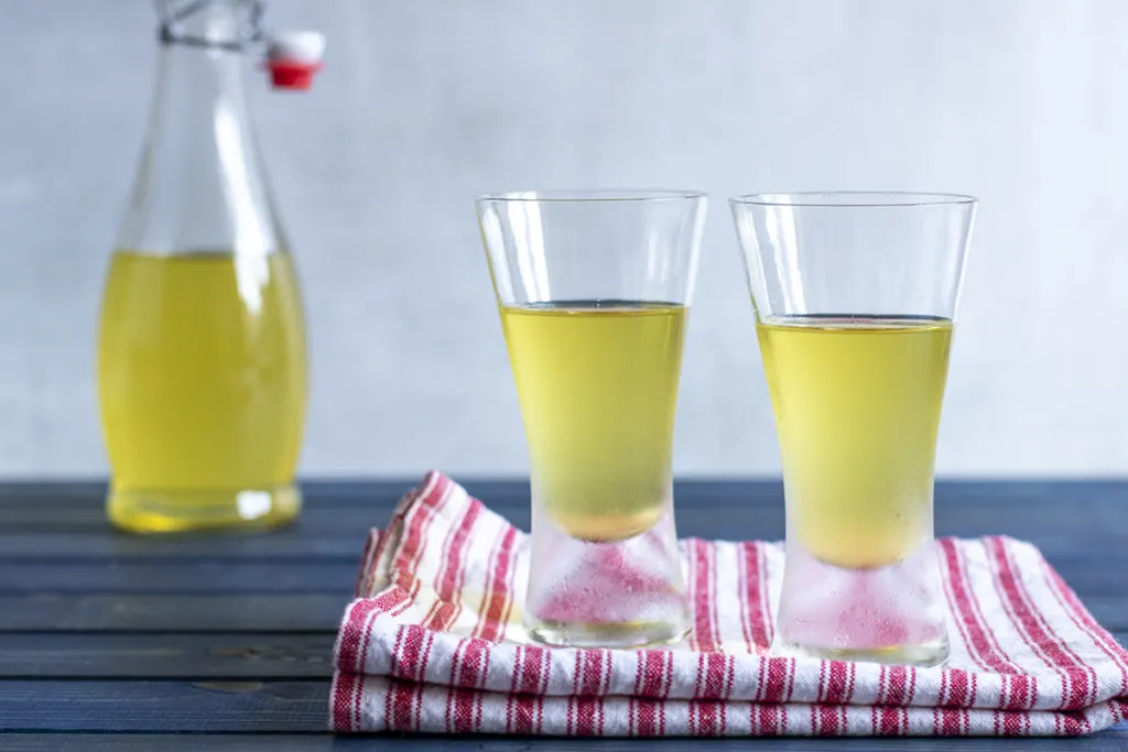 Two glasses of limoncello sit on a folded tea towel. The bottle of limoncello is in the background.