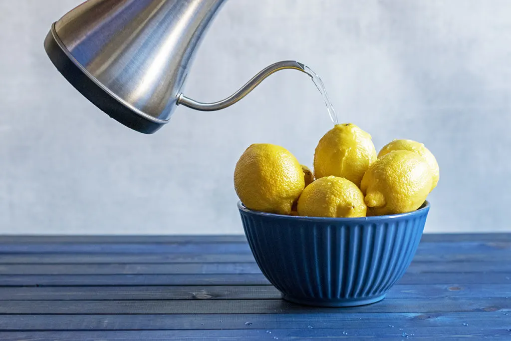 A bowl of lemons on a blue wood table top. Someone is pouring boiling water from a kettle over the lemons.