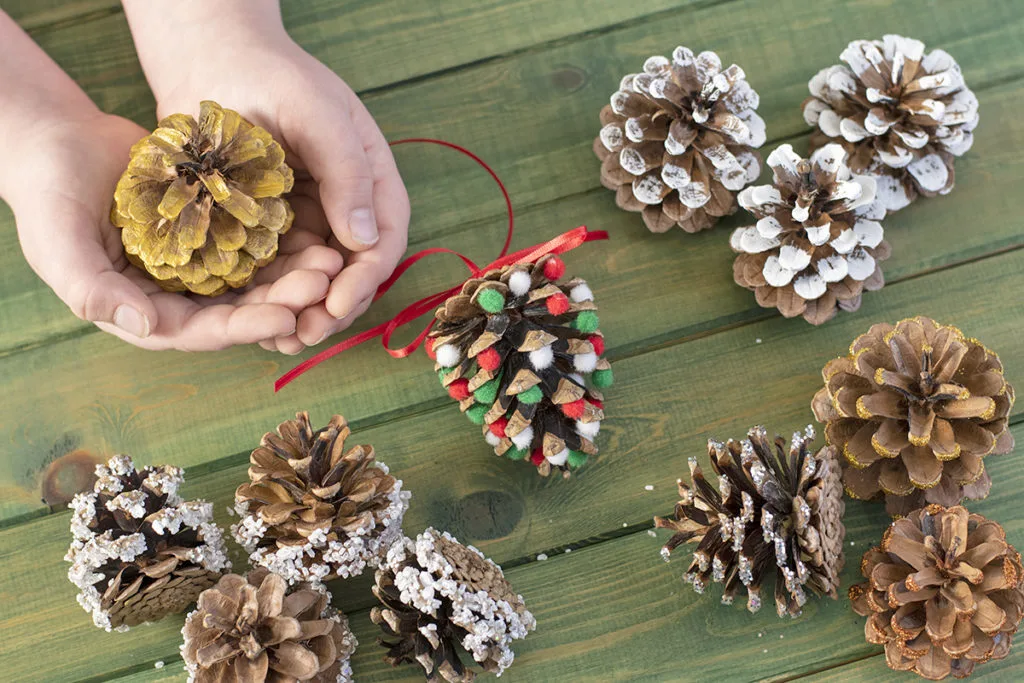 A small child's hands hold a gold-painted pine cone. Several other decorated pine cones are nearby. Some are decorated with glitter, pom-poms, painted to look like snow, and frosted.