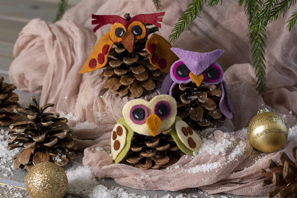 Three owls made out of felt and pine cones are nestled on a piece of cloth with fake snow, Christmas baubles and pine cones.