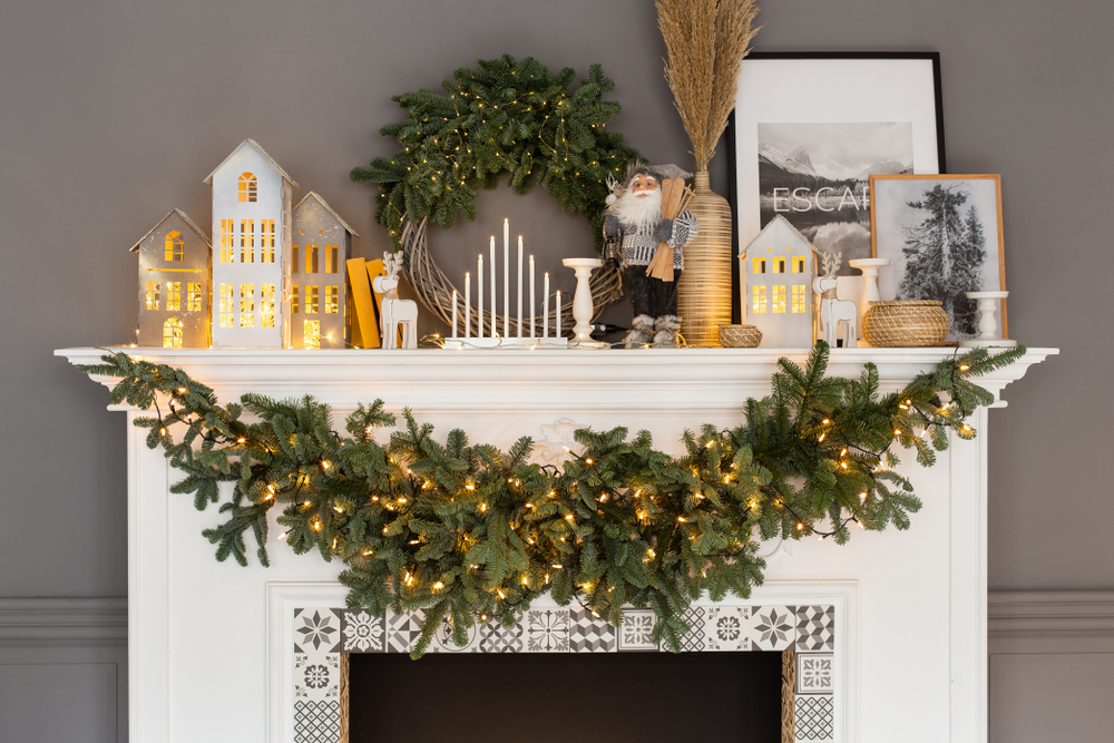 A mantlepiece is decorated with white Christmas decorations. An evergreen garland is hung from the mantlepiece. 