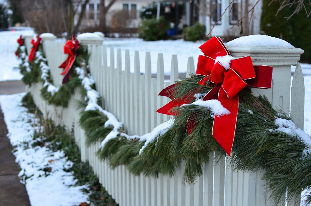 A pine garland hangs on a white picket fence. The garland has snow and red bells on it.