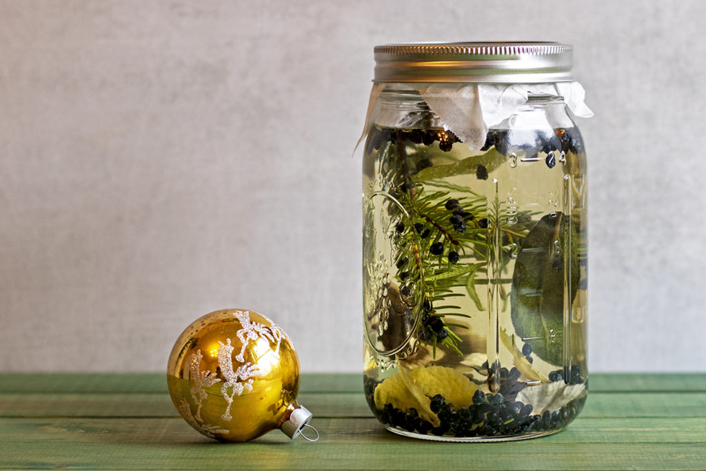 A mason jar filled with botanicals and vodka to make homemade infused gin. A gold Christmas bauble is next to the jar.