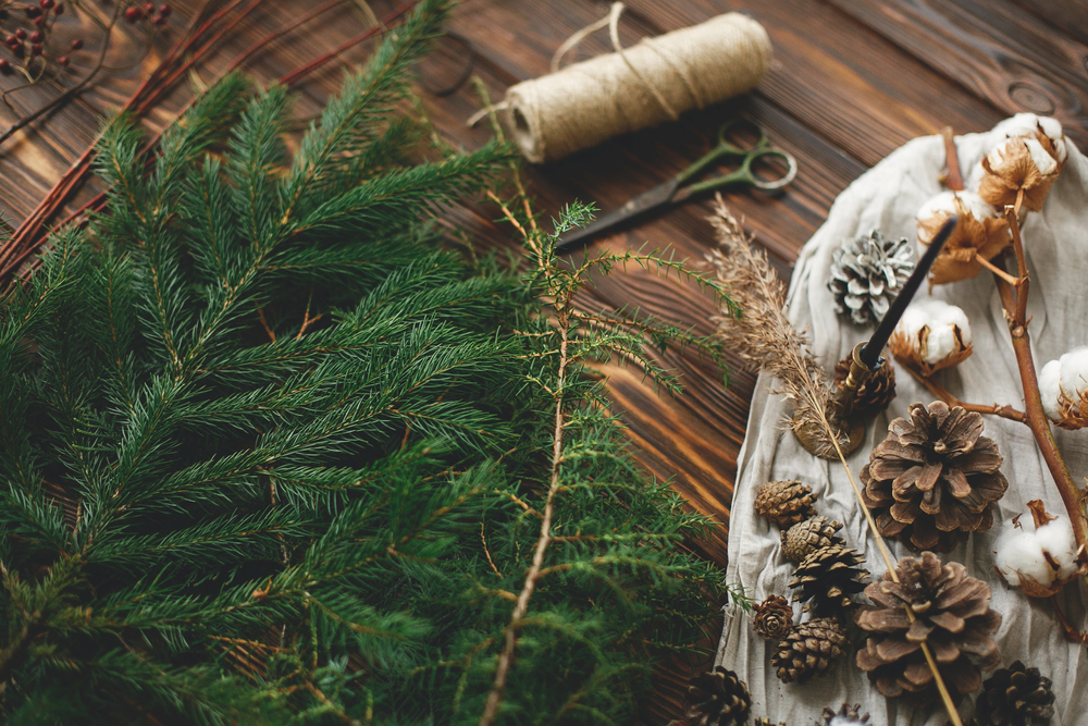 Pine branches, twine and scissors are laid out on a table next to pine cones and cotton.