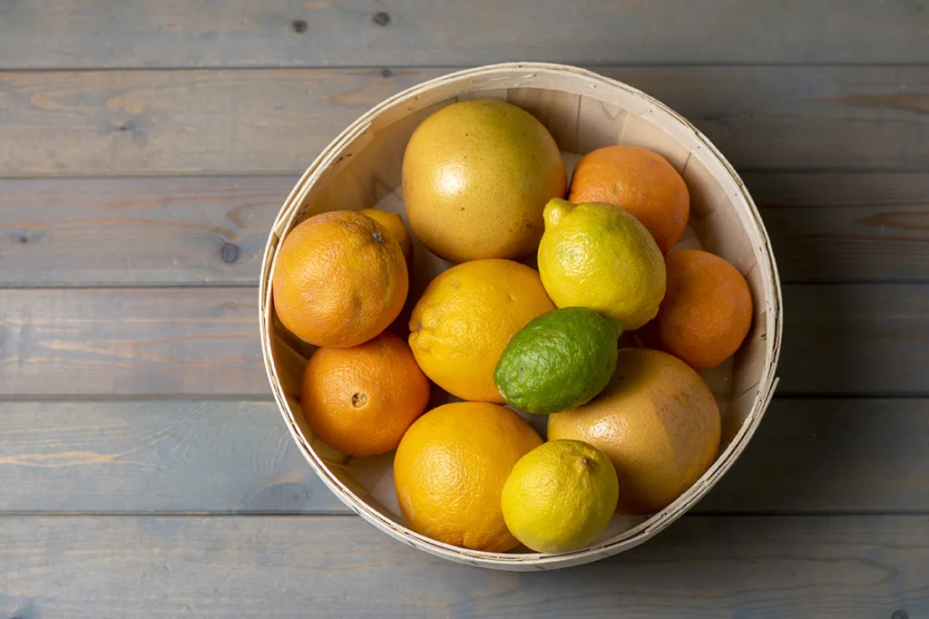A basket of mixed citrus fruit sets on a wooden tabletop.