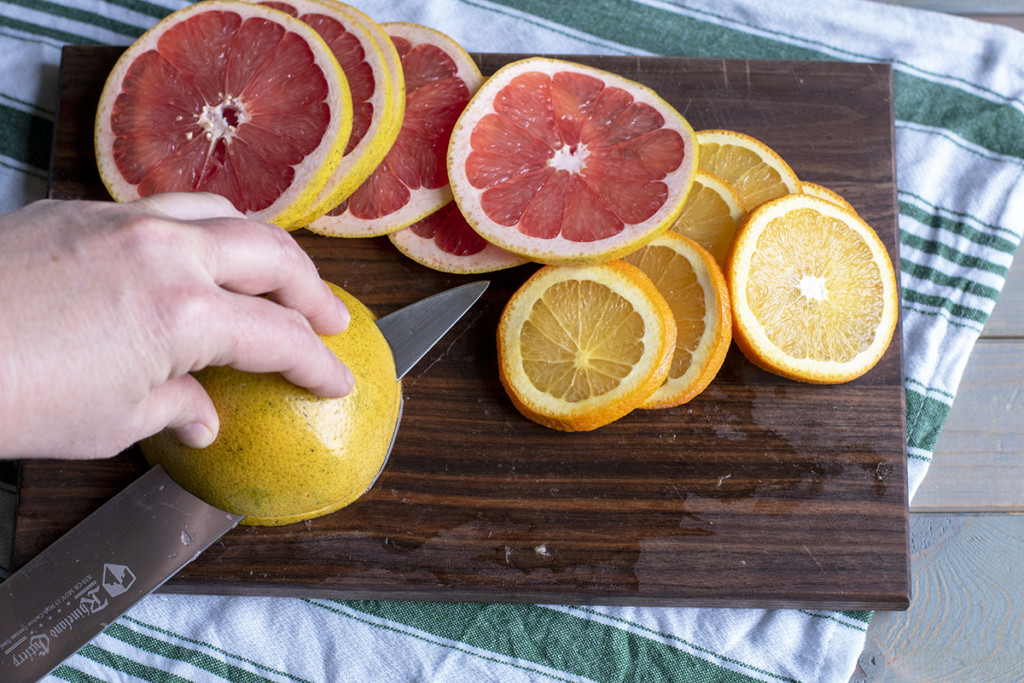 A hand holds a grapefruit while slicing it on a cutting board. There are slices of other citrus fruit nearby.