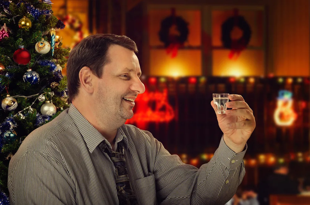 A smiling man holds a shot of vodka, he is sitting in front of a Christmas tree.
