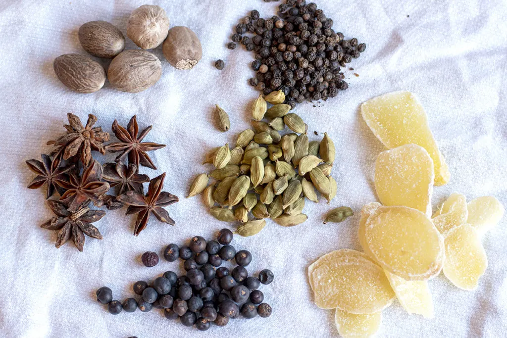 Nutmeg, black peppercorns, ginger slices, cardamom pods, star anise, and junipers grouped together on a white tea towel.